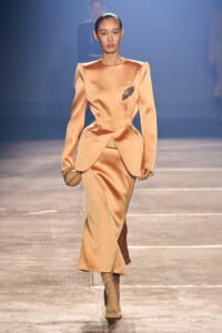Model walks the runway in a peach satin tailored suit with a peplum-like waist and a jeweled brooch, paired with matching skirt and boots.