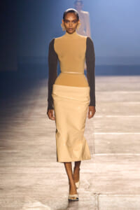 Model on a runway wearing a tan sleeveless turtleneck top with dark brown sleeves and a glossy beige midi skirt, with large gold earrings.