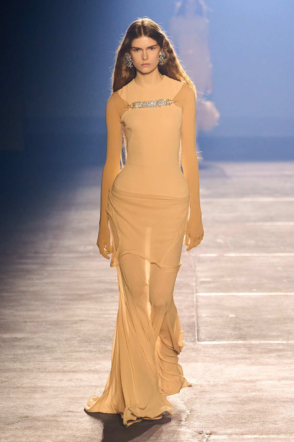 Model walks the runway in a long, beige, form-fitting gown with a jeweled chest band and draped, flowing skirt.