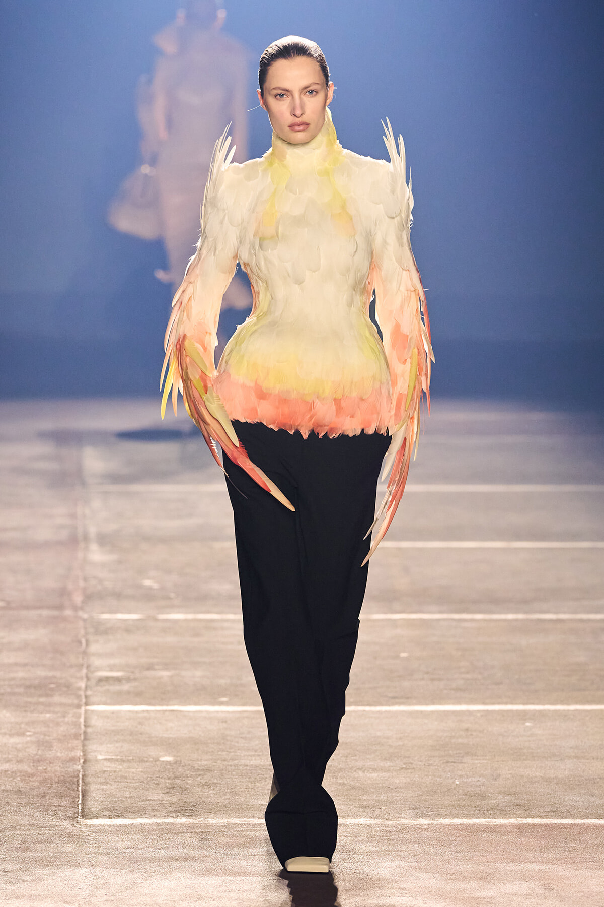 Model walking a runway in a feathered, bird-inspired top with white, yellow and pink hues, paired with black trousers and white shoes.