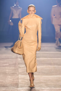 Model walks the runway in a tan sculptural midi dress with an oversized collar, carrying a matching beige handbag