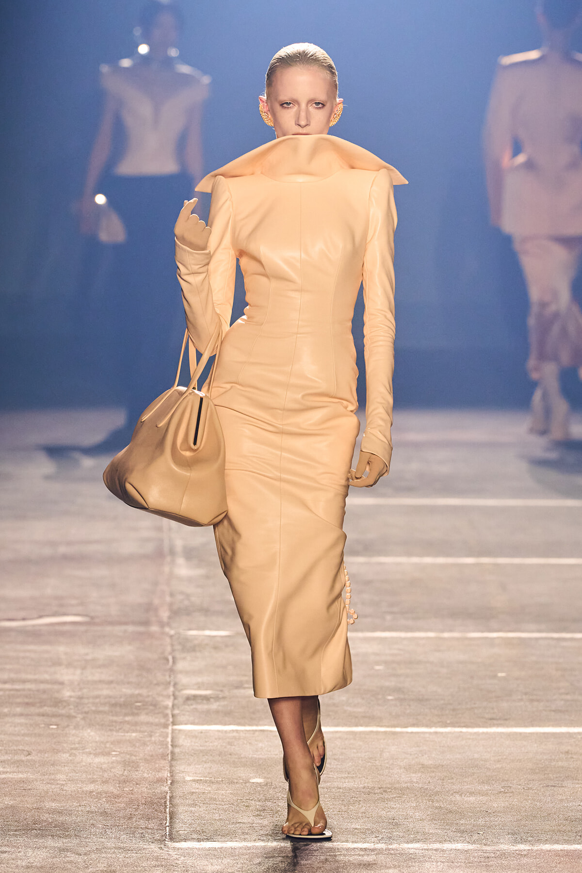 Model walks the runway in a tan sculptural midi dress with an oversized collar, carrying a matching beige handbag