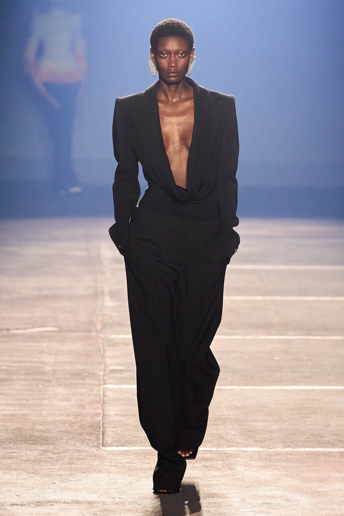 Female model walks a runway in a black tailored suit with a plunging draped blazer and wide-leg pants, accessorized with bold statement earrings.