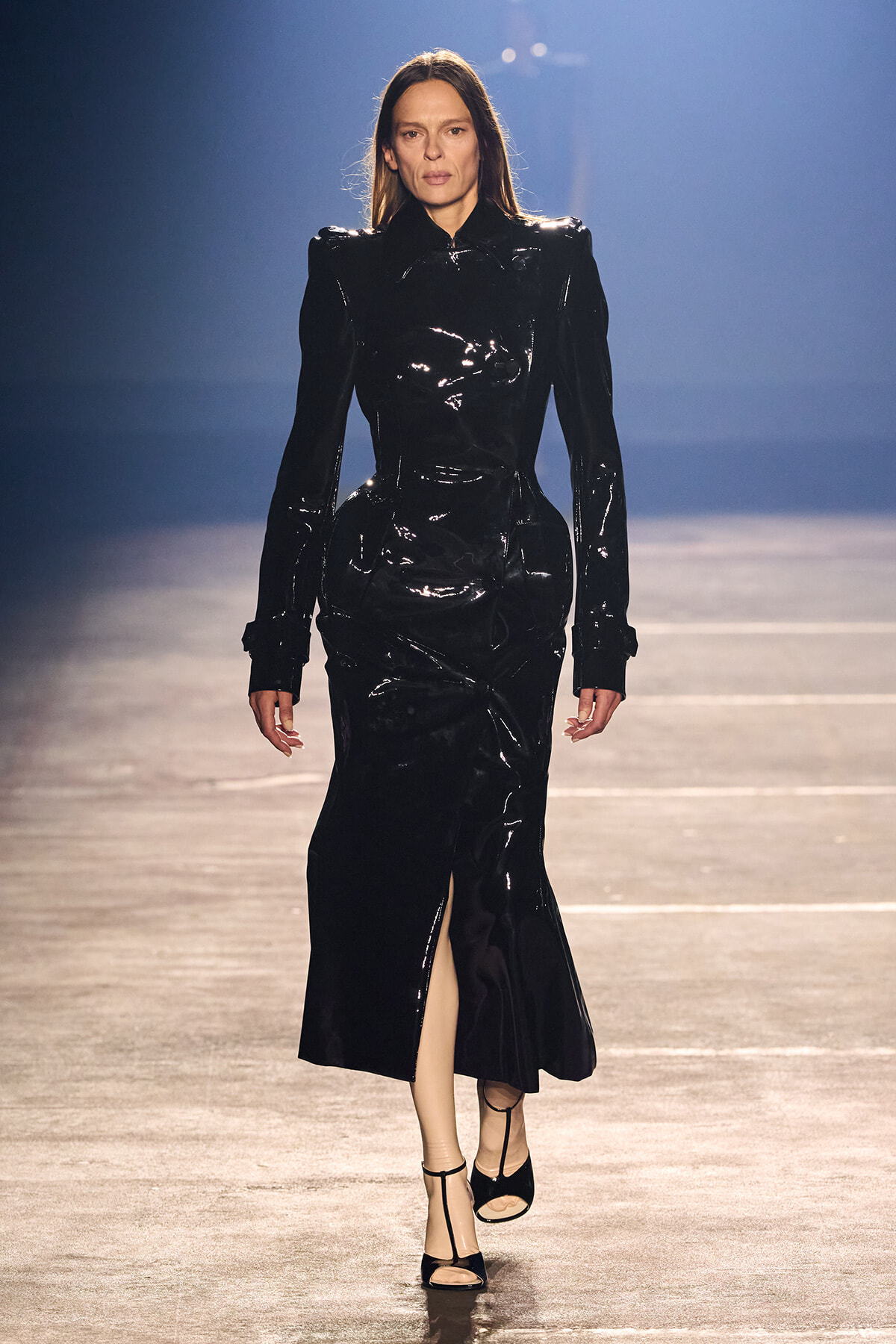 Runway model wearing a long, glossy black patent dress with structured shoulders, high collar, and a front slit, paired with black ankle-strap heels.