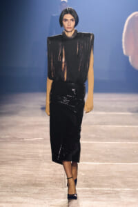 Model walking a fashion runway in a black fringe blouse with mustard sleeves and a shiny black patent skirt, dark ankle-strap heels.