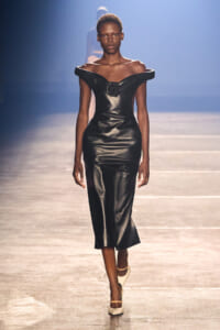 Model walks the runway in a sleek black off‑shoulder leather midi dress with a central rose detail and cream heels.