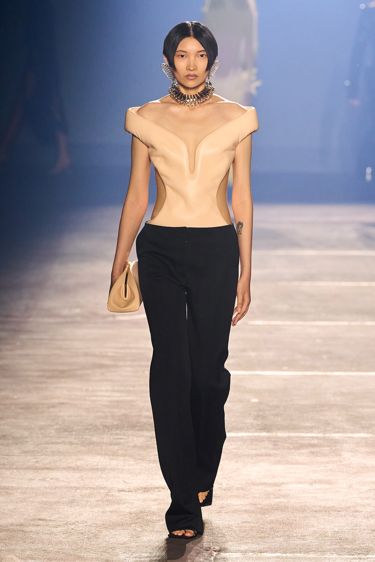 Runway model in a sculptural nude top with black wide-leg pants, carrying a beige clutch and wearing a bold choker necklace.