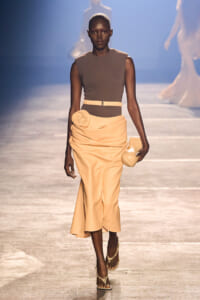 Runway model wearing a taupe sleeveless top with a peach draped skirt, belt, flower detail at the waist, and a matching clutch.