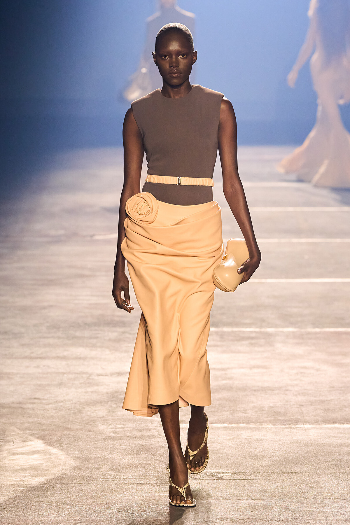 Runway model wearing a taupe sleeveless top with a peach draped skirt, belt, flower detail at the waist, and a matching clutch.