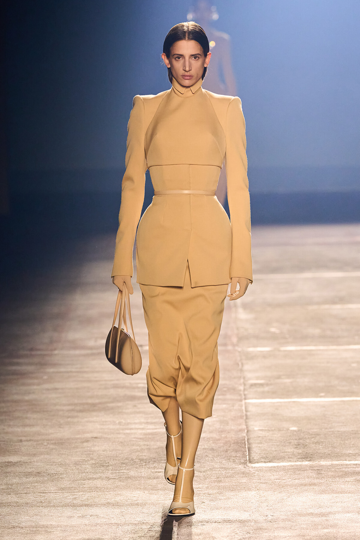 Model walks the runway in a monochrome beige tailored suit with structured shoulders, high collar, and a matching belt, carrying a beige handbag.