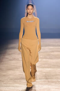 Model walks the runway in a tan, long-sleeve top with crystal embellishment at the chest and wide-leg satin pants; large floral earrings.