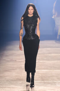 Model walking a runway in a black, sculpted corset with ornate detailing and a long black skirt, paired with black ankle boots.