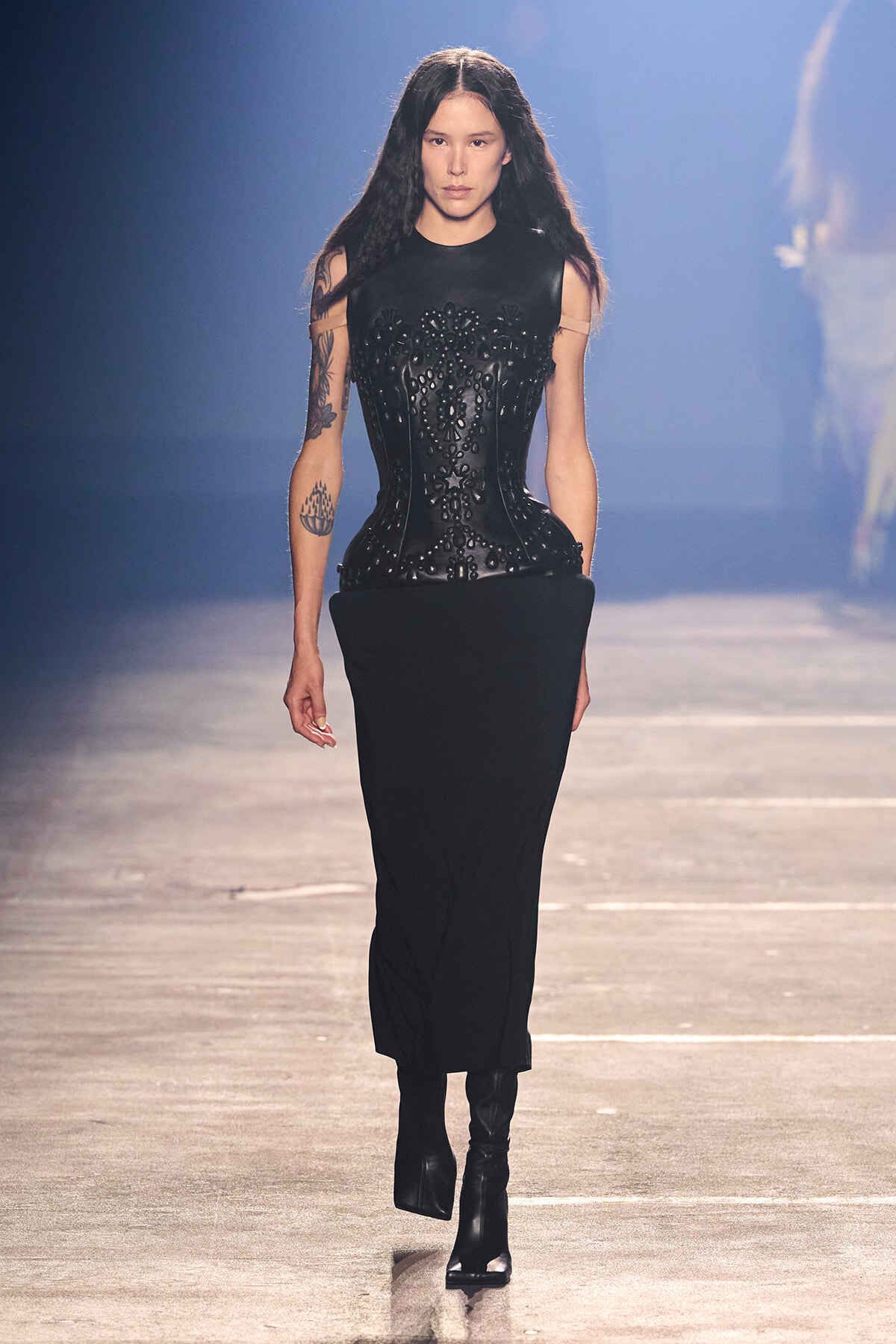 Model walking a runway in a black, sculpted corset with ornate detailing and a long black skirt, paired with black ankle boots.