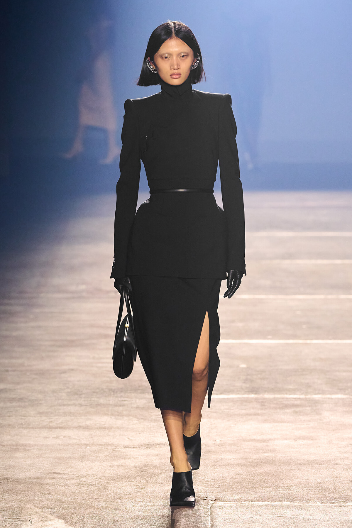 Model on a runway wearing an all-black, structured ensemble: high-neck top with padded shoulders, belted waist, and a midi skirt with a thigh-high slit, carrying a small black handbag and wearing gloves.