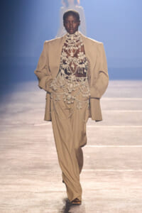 Runway model walks in a loose beige suit over a sheer beaded dress with layered white jewelry on a fashion catwalk.