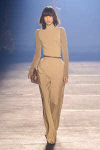 Model walking a beige-toned runway in a cream turtleneck with long beige sleeves and high-waisted wide-leg pants, belt, and small brown handbag.
