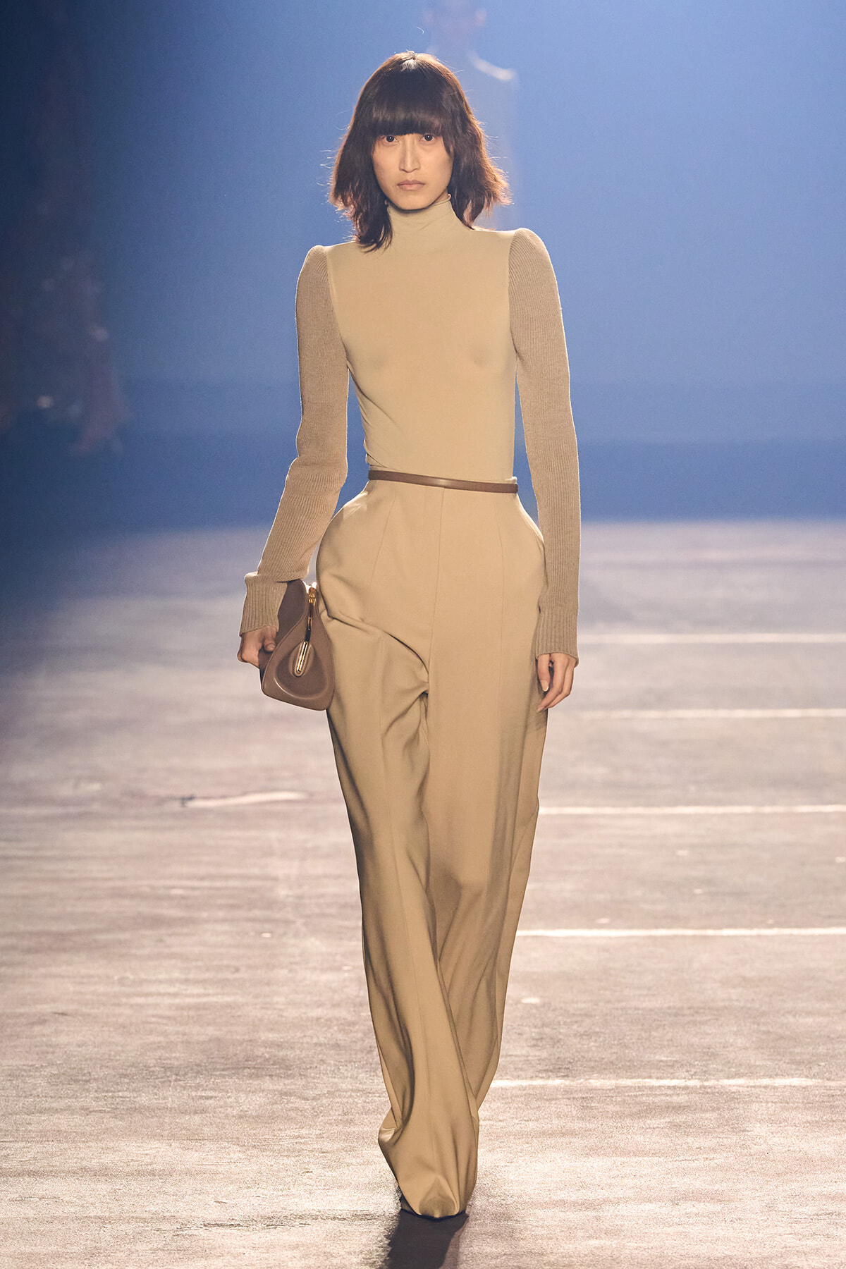 Model walking a beige-toned runway in a cream turtleneck with long beige sleeves and high-waisted wide-leg pants, belt, and small brown handbag.