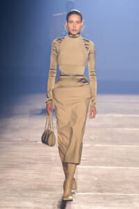 Model on a runway wearing a beige, high-neck, long-sleeve dress with matching boots and a small beige handbag.