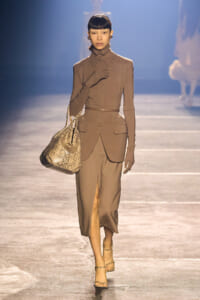 Model walking runway in a taupe suit with a belted waist, matching gloves, and a snakeskin tote bag, wearing strappy heels.