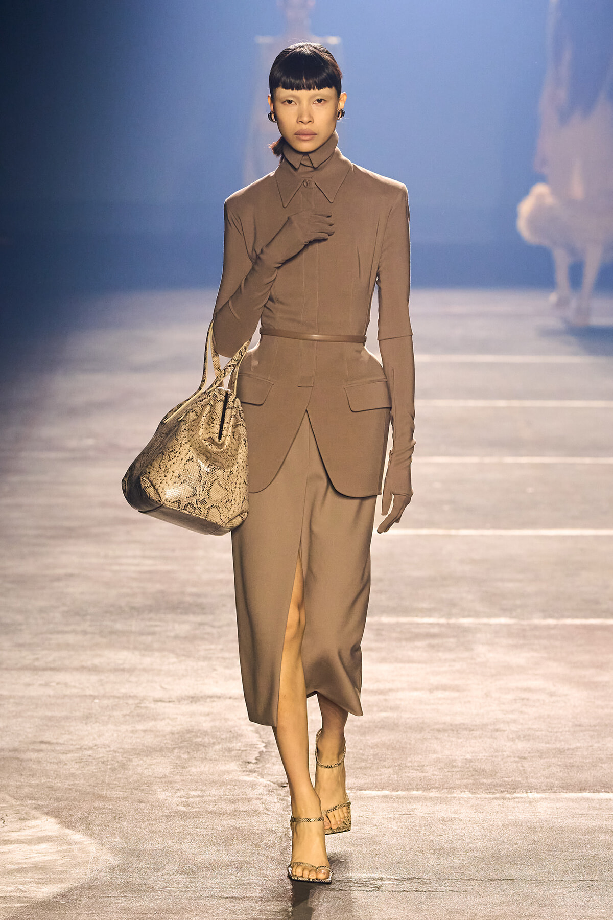 Model walking runway in a taupe suit with a belted waist, matching gloves, and a snakeskin tote bag, wearing strappy heels.