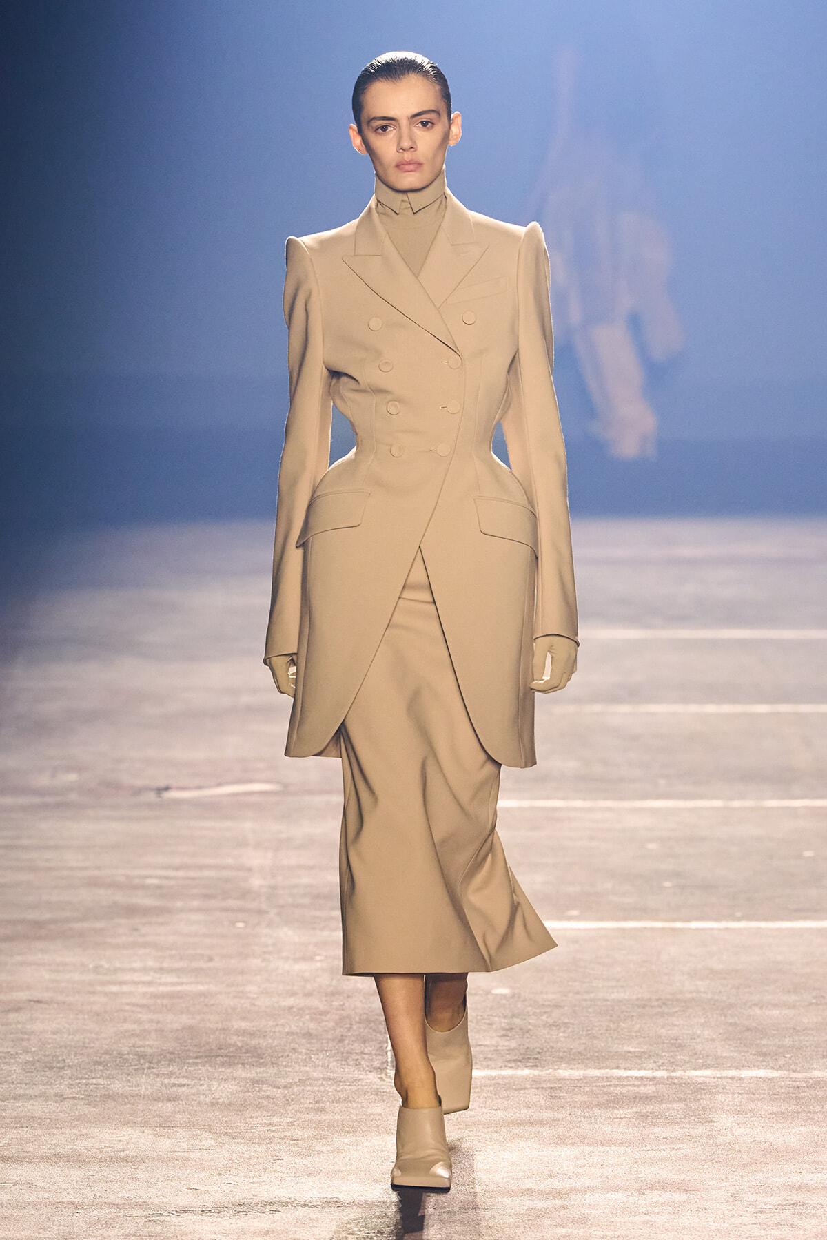 Model walks the runway in a beige, tailored, double‑breasted coat-dress with a front wrap, matching gloves, and beige heeled boots.