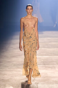 Runway model walks forward in a nude-toned, star-sequined draped gown that exposes her shoulders and torso.