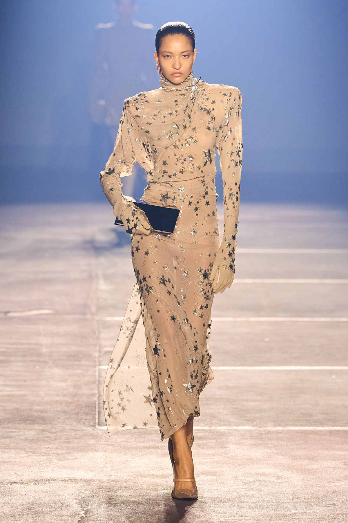 Beige, full-length gown with silver star embellishments, high neck, long sleeves, matching gloves, and a black clutch as the model walks a runway.