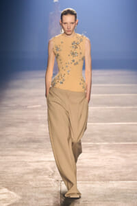 Model walking a runway in a beige sleeveless top with silver floral embroidery and wide tan trousers on a blue-lit stage.