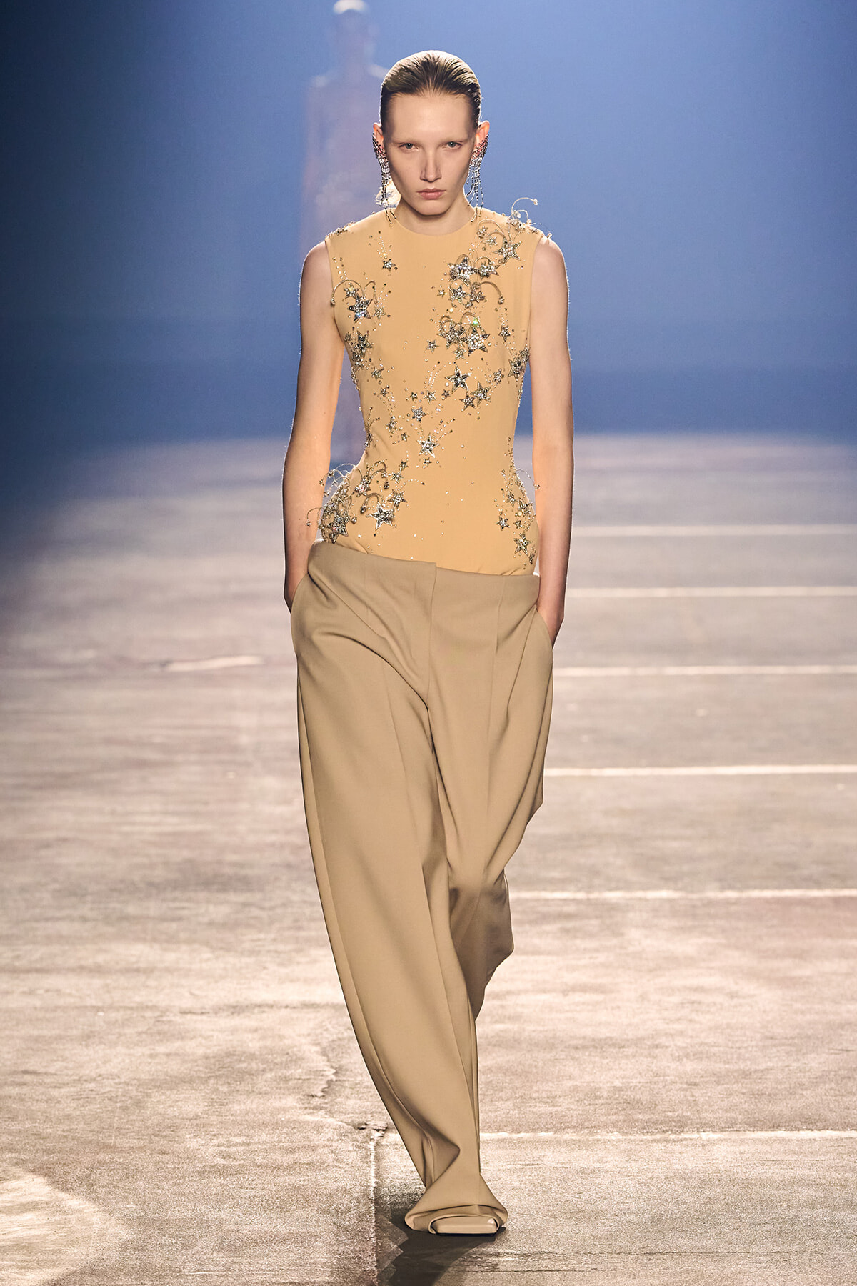 Model walking a runway in a beige sleeveless top with silver floral embroidery and wide tan trousers on a blue-lit stage.