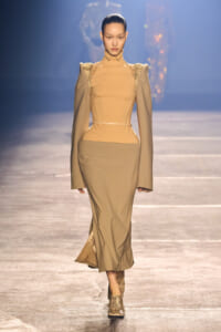 Model walking a runway in a tan, high-neck, fitted dress with exaggerated shoulders and long sleeves, paired with a matching long skirt and snakeskin ankle boots.