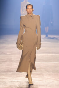 Model walks the runway in a beige double-breasted trench coat with structured shoulders, tan boots, and a small snake-print clutch.