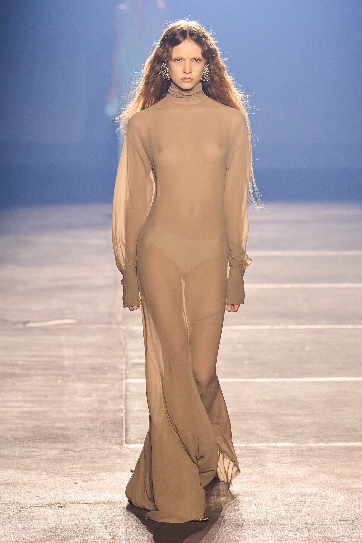 Model walking a fashion show runway in a full-body sheer nude bodysuit with long sleeves and oversized cuffs, beige tones, dramatic earrings начали.