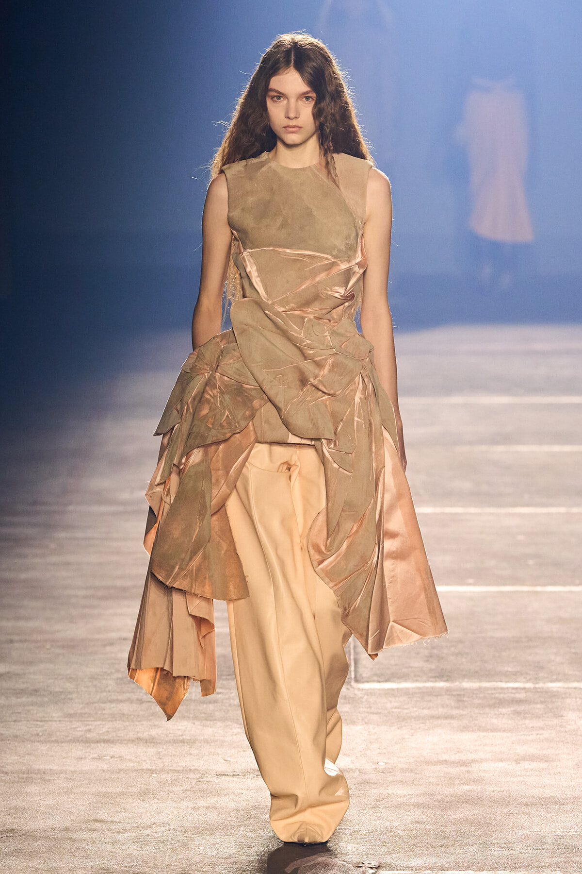 Model walking the runway in a sleeveless beige gown with sculptural draped panels and peach satin underlayers.