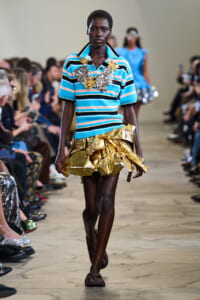 Model walks the runway in a turquoise striped polo with a large gold metallic ruffled skirt and statement gold floral jewelry on the chest area.
