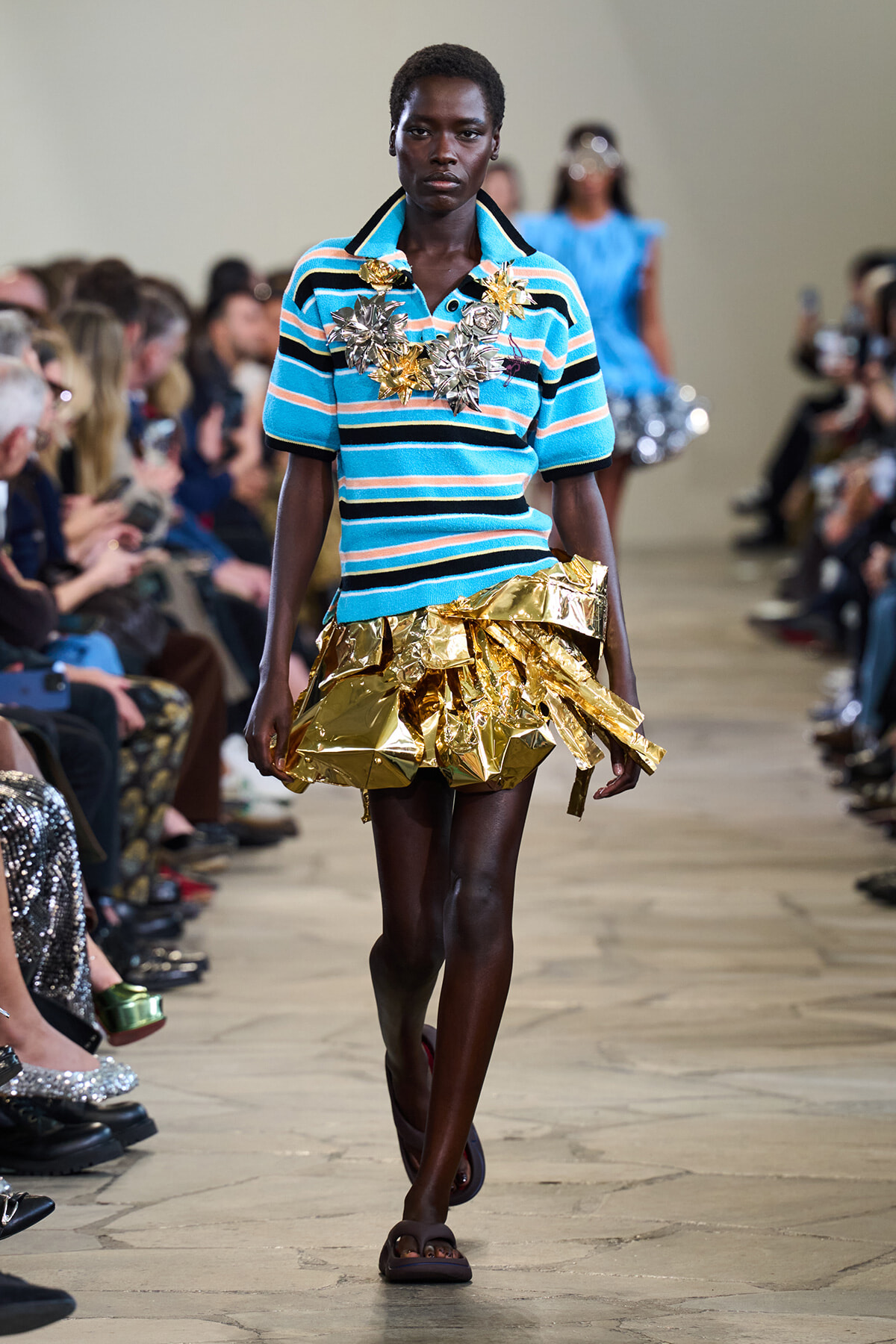 Model walks the runway in a turquoise striped polo with a large gold metallic ruffled skirt and statement gold floral jewelry on the chest area.