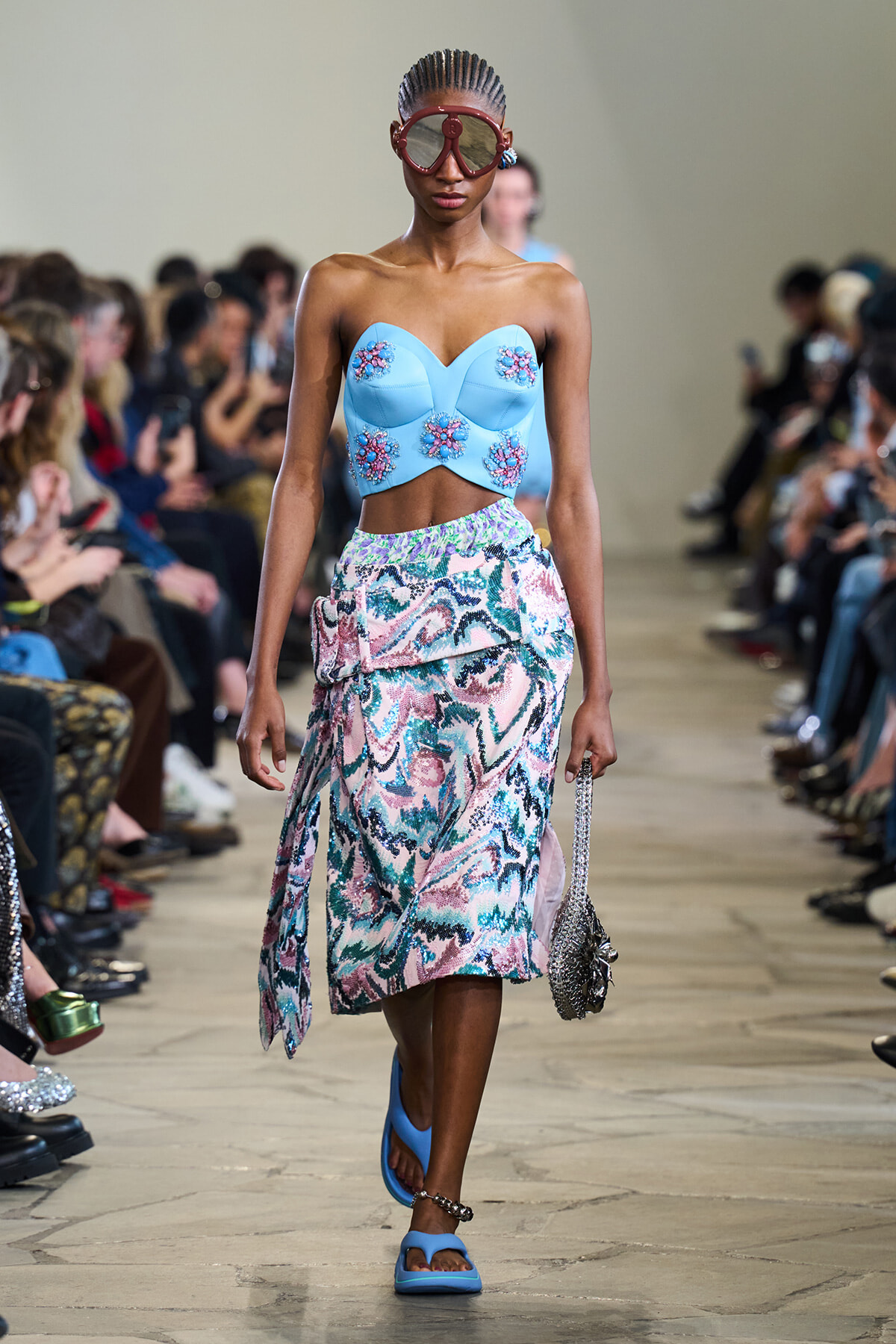 Model walking a runway in a pale blue strapless top with floral embroidery and a colorful patterned midi skirt.