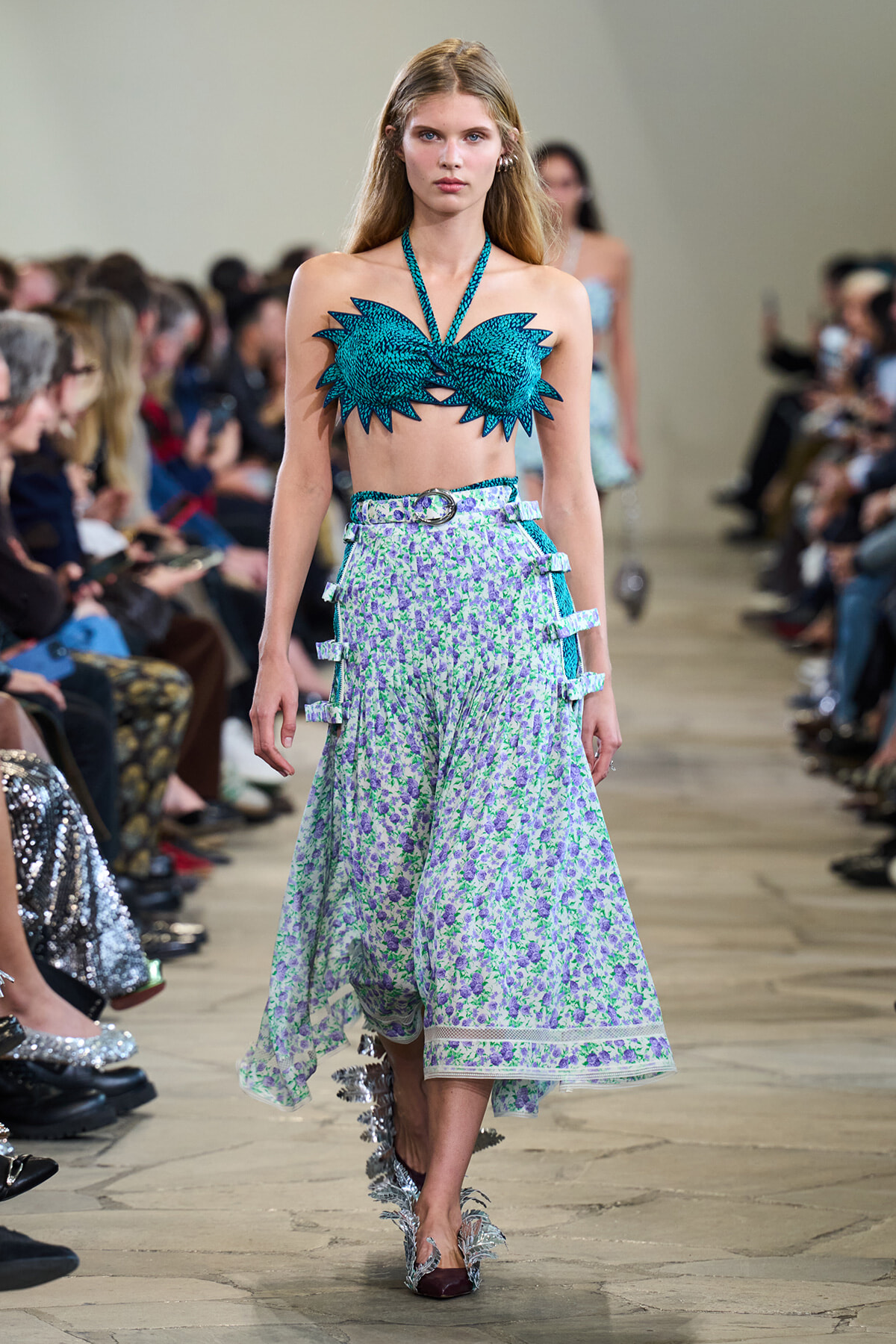 Model walks the runway in a turquoise spiked bandeau top and a light floral midi skirt with a belt and side strap details.