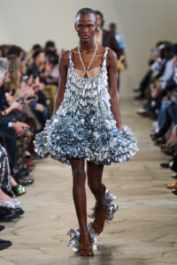Runway model wearing a silver, ruffled, leaf-like dress with a V-neck and halter straps, walking the catwalk as photographers line the audience behind her.