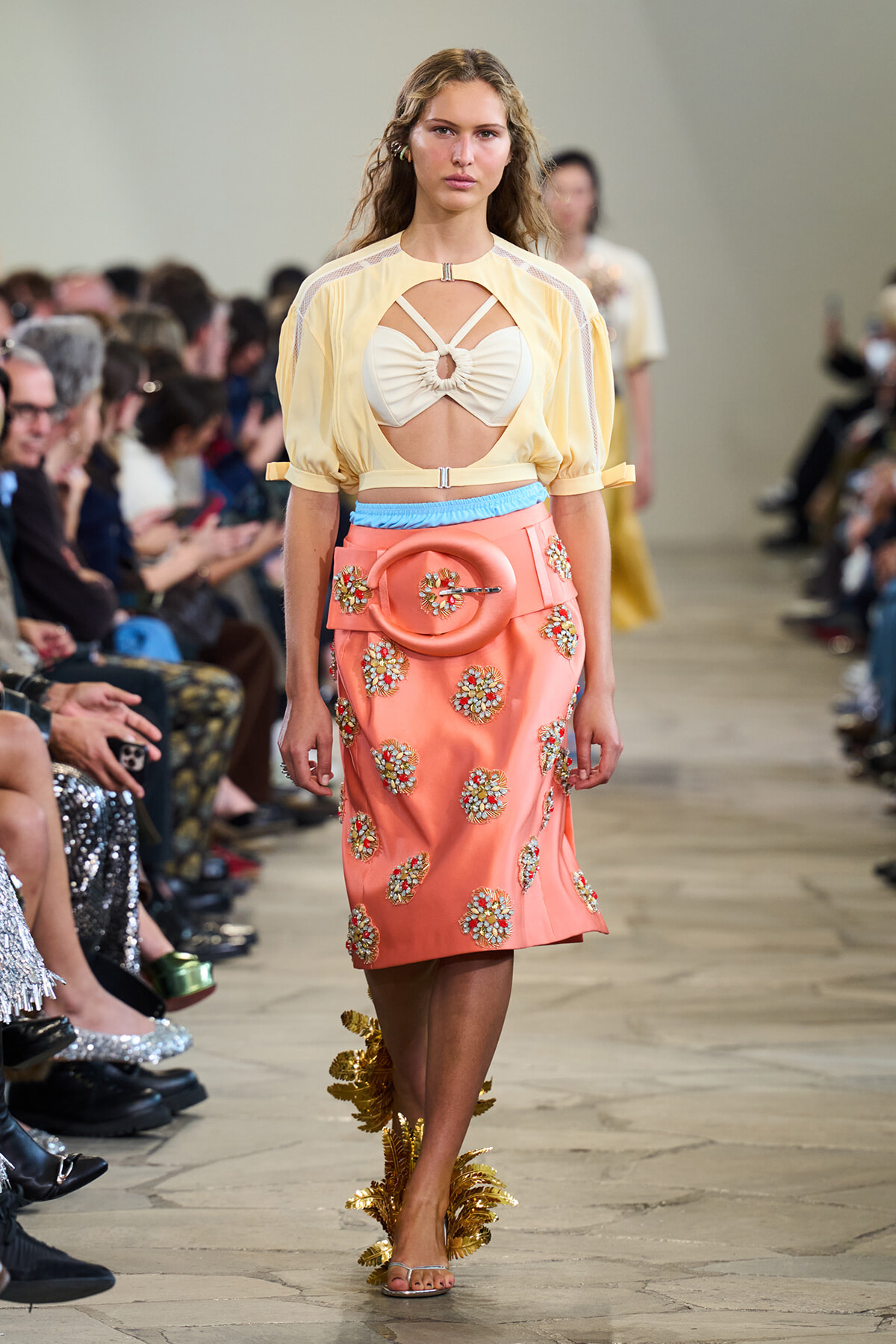 Model walks the runway in a pale yellow sheer top with a cutout bodice and a peach embellished skirt with floral appliqué and a circular belt centerpiece.