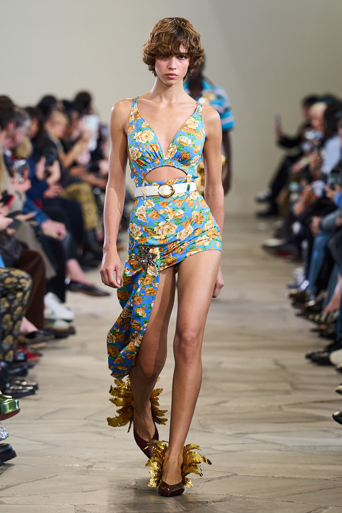 Model on a runway wearing a turquoise floral two-piece outfit with a cutout at the midriff, white belt, and a wrap skirt with gold leaf-embellished heels.