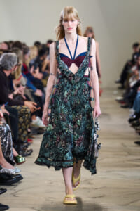Model walking a runway in a tropical-green printed dress with blue halter straps and burgundy top, wearing yellow sole sandals.
