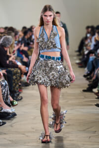 Female model walks a runway in a blue-and-white knit crop top and a short metallic-flower skirt with a blue belt; oversized silver floral embellishments dominate the outfit.