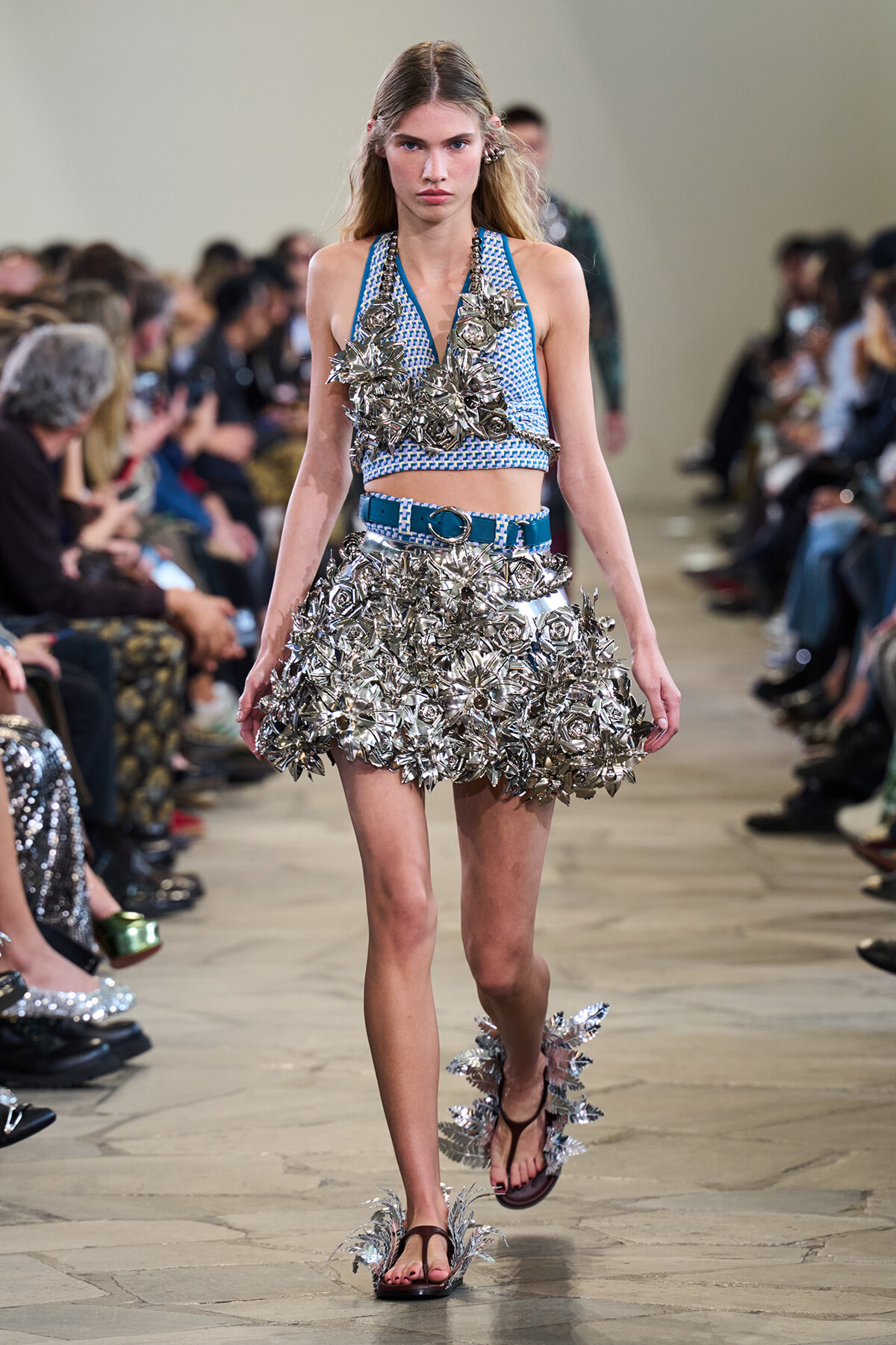 Female model walks a runway in a blue-and-white knit crop top and a short metallic-flower skirt with a blue belt; oversized silver floral embellishments dominate the outfit.