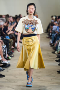 Runway model wearing a cream top with jeweled palm-tree appliqués over a blue-green cutout, and a high-waisted yellow skirt with a large circular belt buckle; blue sandals visible.