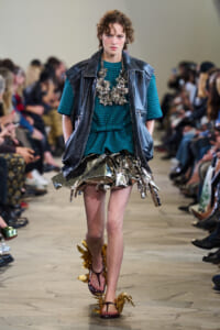 Model walks the runway in a teal knit top under a black leather vest, accessorized with a bold metallic flower necklace and a crumpled metallic skirt, finished with gold-leaf sandals.