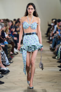 Model walking a fashion runway in a lavender floral two-piece with ruffled skirt and halter top, silver heels, and a metallic handbag.