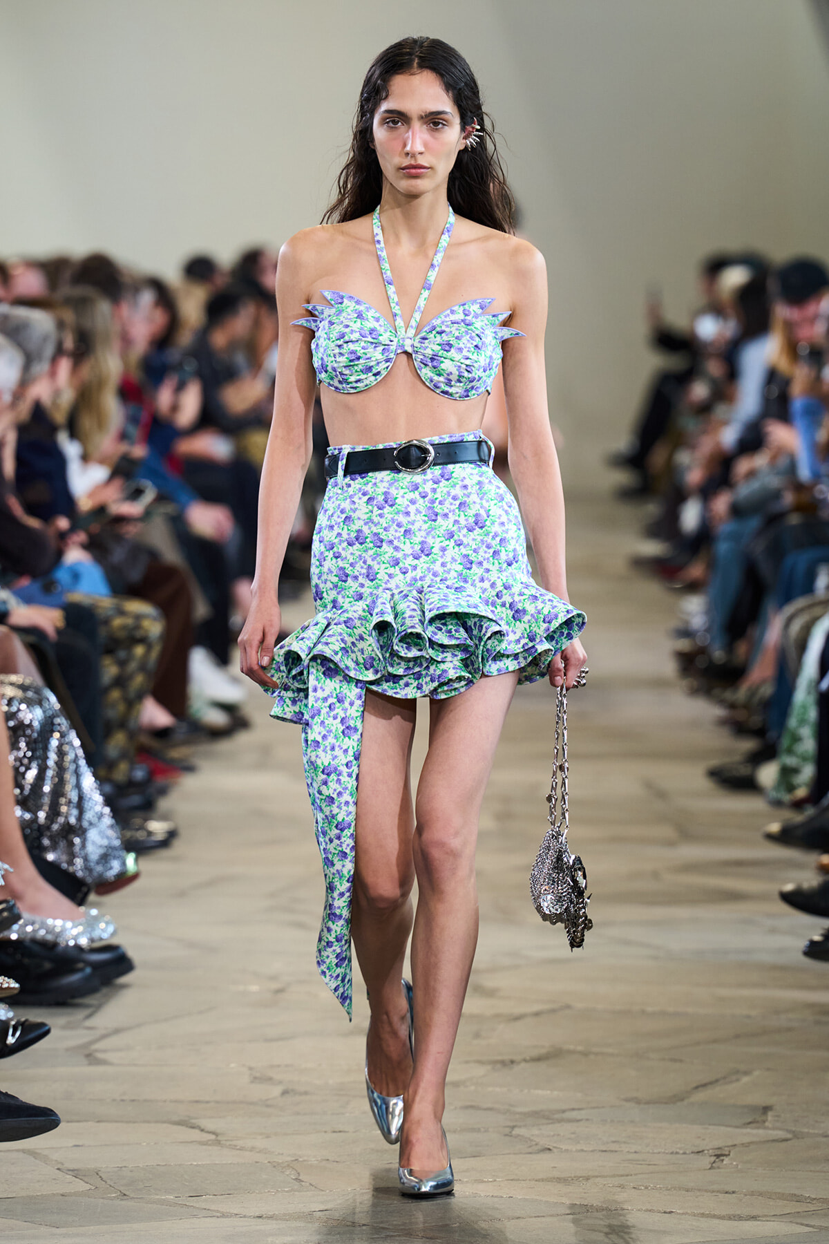 Model walking a fashion runway in a lavender floral two-piece with ruffled skirt and halter top, silver heels, and a metallic handbag.