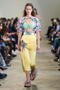Model walking a runway in a pastel outfit: mint green cutout top with crisscross straps and a pale yellow pencil skirt with a metallic floral embellishment.