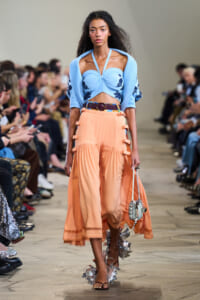 Runway model wearing a light blue cropped top with matching shawl and peach high-waisted pleated skirt, walking the catwalk with audience in background.