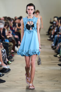Model on a runway wearing a light blue dress with large blue floral appliqués around the neckline and hem, black bodice underneath, and floral sandals; audience blurred in background.
